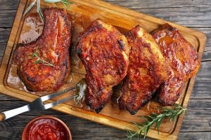 Beautifully caramelized pork chops, on a wooden cutting board with rosemary, barbecue utensils, and a bowl of sauce